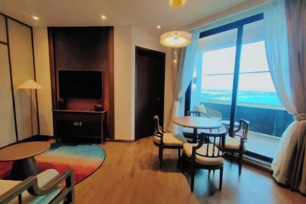 Habitación de hotel con un ventanal grande, un televisor de pantalla plana, una mesa redonda con sillas, un sofá pequeño y una alfombra colorida.