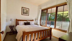 Ein Schlafzimmer mit einem hölzernen Bett, einem Nachttisch, einem gerahmten Bild an der Wand und einem großen Fenster mit Vorhängen.