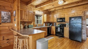 A wooden cabin kitchen with wooden cabinets, a granite countertop, a black refrigerator, a microwave, a stove, and a sink. - Cozy Log Cabin - Hot Tub, Games, & Mountain Views (Sevierville)