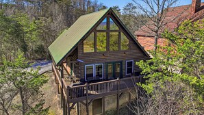 A wooden cabin with a green roof, large windows, and a deck. - Cozy Log Cabin - Hot Tub, Games, & Mountain Views (Sevierville)