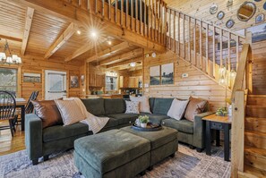 Interior acolhedor de uma cabana com escada de madeira, um amplo sofá modular e uma área de jantar.
