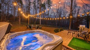 Un jacuzzi en una terraza de madera con sillas y una parrilla, rodeado de árboles y luces colgantes.