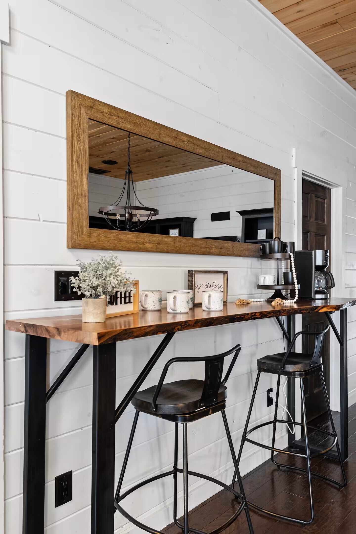 A kitchen with a wooden bar counter, two black bar stools, a large mirror, and a hanging light fixture.