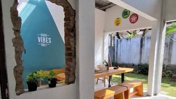 Ein überdachter Außenbereich mit Holzbänken und einem Tisch, eine Wand mit dem Schriftzug 'VIBES ONLY' sowie ein kleiner Garten mit Topfpflanzen.