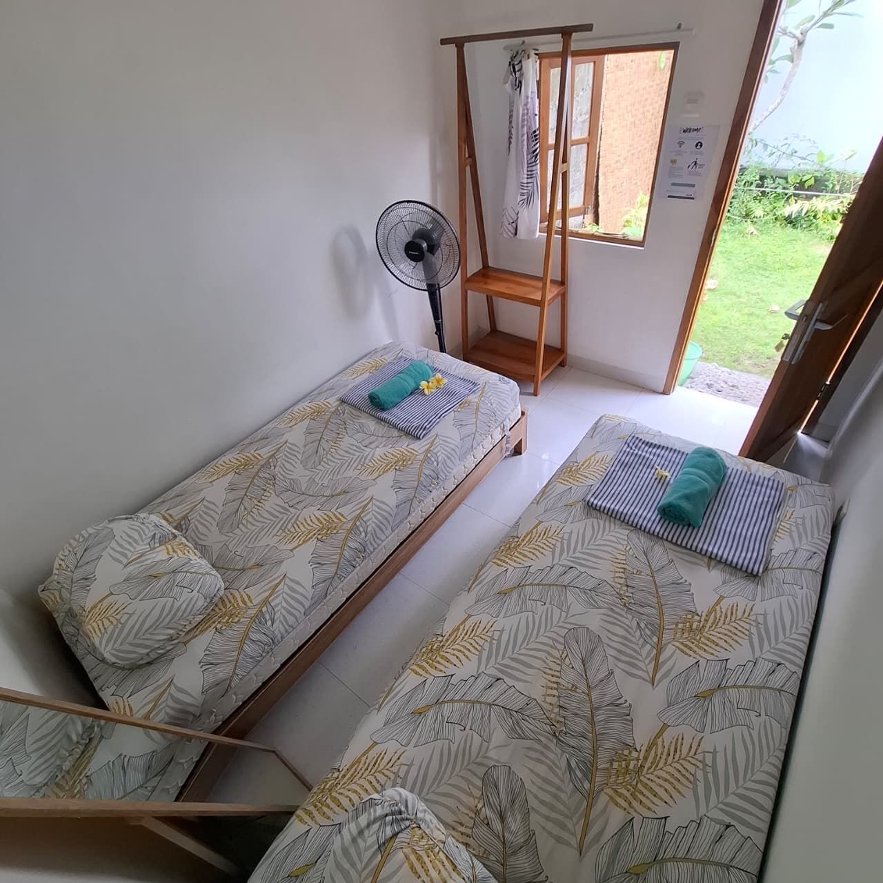 A room with two single beds, a standing fan, a wooden shelf, and a window with a view of greenery.