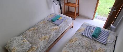 A room with two single beds, a standing fan, a wooden shelf, and a window with a view of greenery.