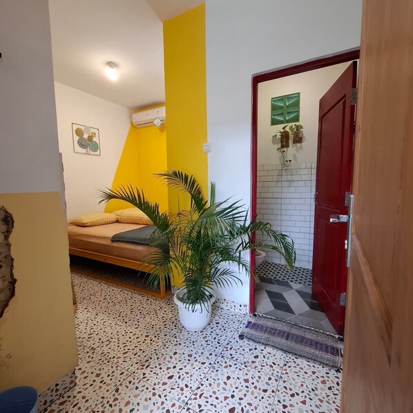 A small, brightly colored room with a bed, a potted plant, and a red door leading to a bathroom with a tiled floor. - The Backyard Hostel (Yogyakarta)