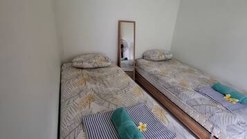 Un dormitorio con dos camas individuales, cada una con una colcha estampada con hojas y una almohada. Un espejo cuelga en la pared, y se ve un ventilador al fondo.