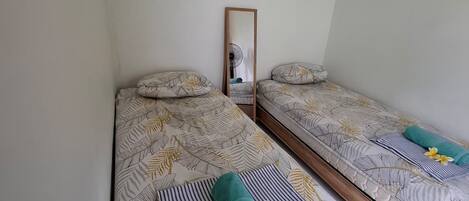 A bedroom with two single beds, each with a leaf-patterned bedspread and a pillow. A mirror hangs on the wall, and a fan is visible in the background.