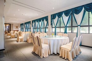Une salle de banquet avec des tables rondes et des chaises, de grandes fenêtres ornées de rideaux bleus et un sol recouvert de moquette.