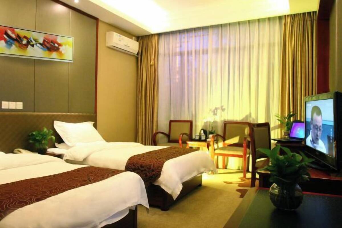 Ein Hotelzimmer mit zwei Betten, einem Fernseher, einem Schreibtisch mit Stuhl und einer Pflanze.