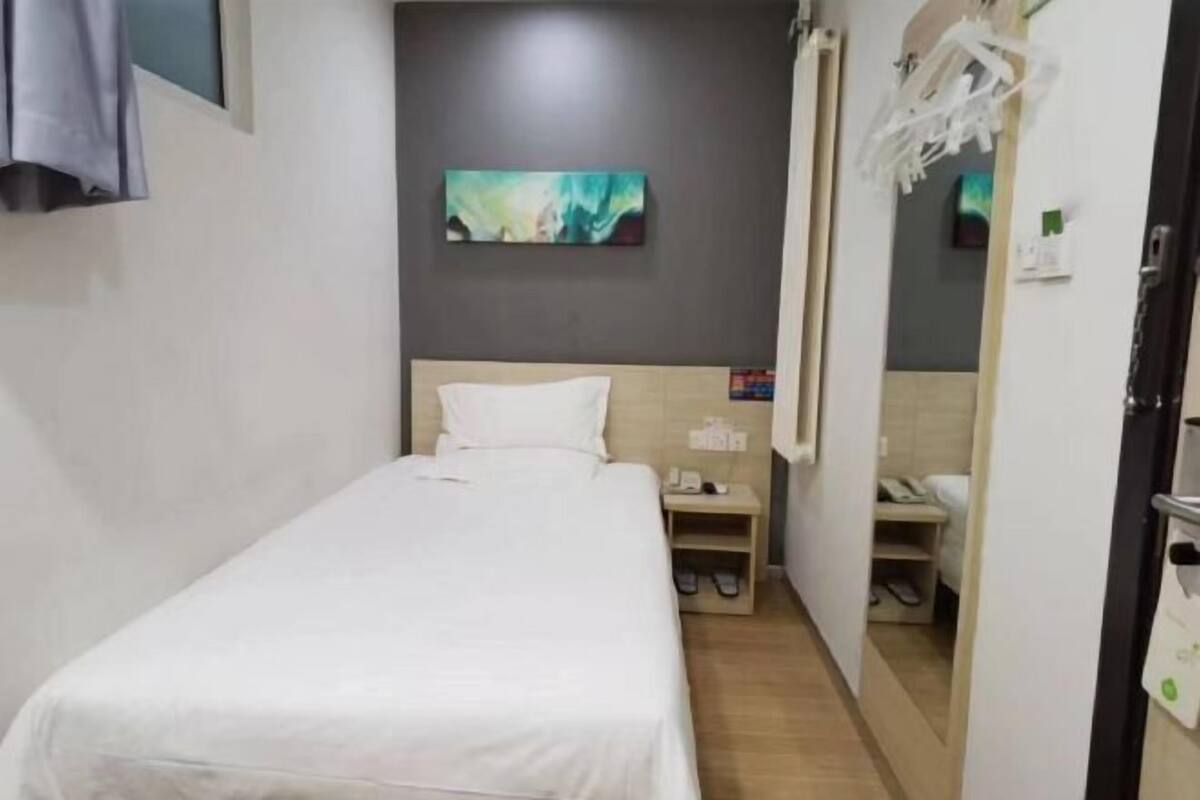 A single bed with a white bedspread, a nightstand with a phone and slippers, a painting on the wall, and a door with a towel rack.