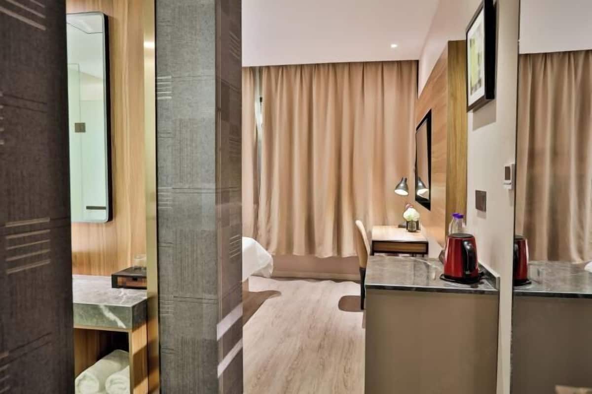 A hotel room with a bed, a desk, a chair, a nightstand, a lamp, and a red kettle.