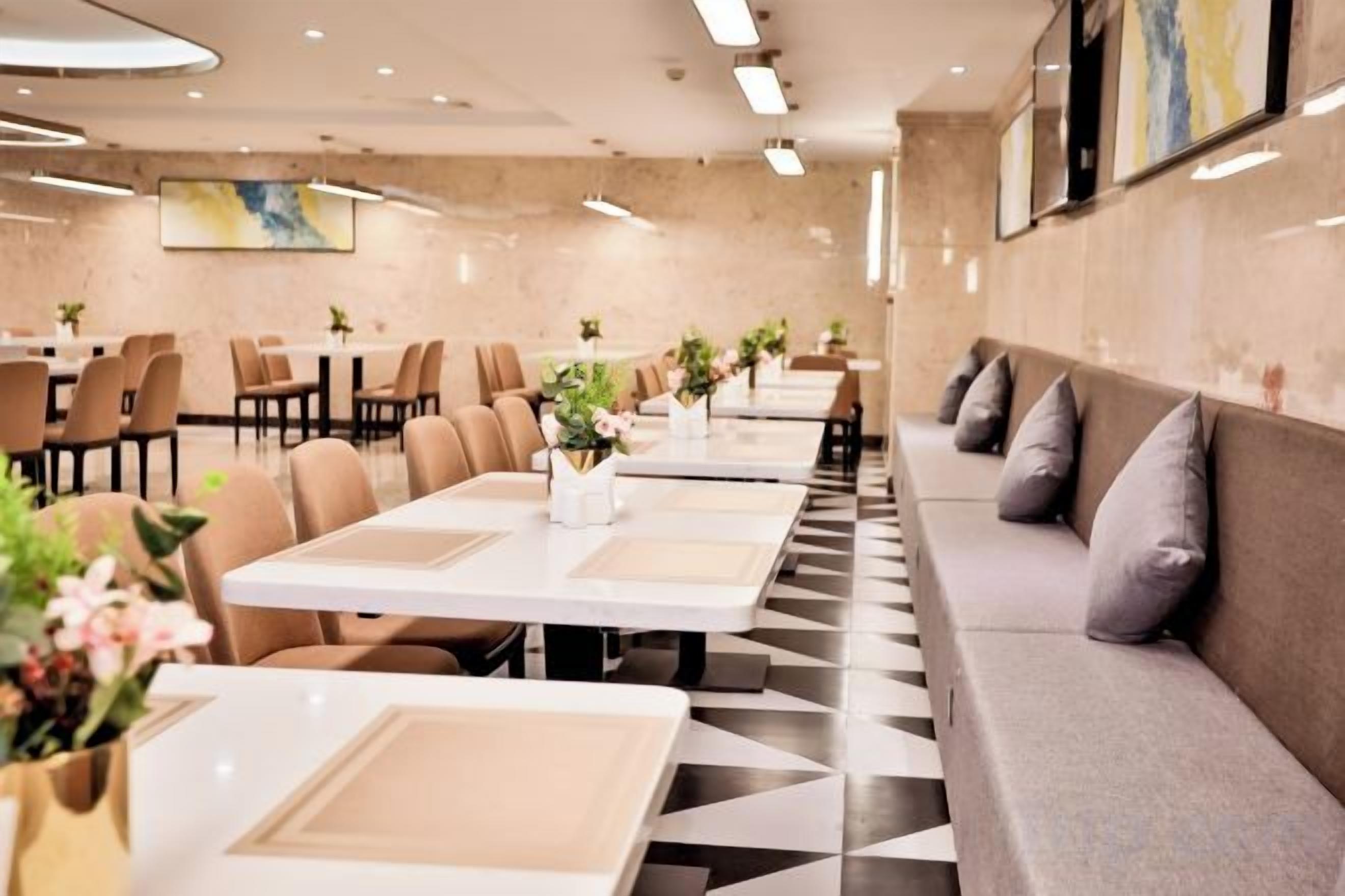 Área de refeições de hotel com mesas compridas, assentos acolchoados e plantas decorativas.