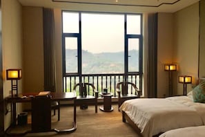 A hotel room with a large window, a bed, a desk with a chair, and two bedside lamps.