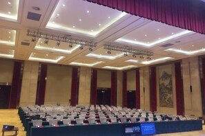 Un salón de conferencias con filas de mesas y sillas, un escenario con equipo de iluminación y un gran mural en la pared.
