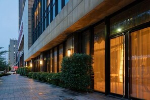 Exterior moderno de un hotel con amplios ventanales, un camino pavimentado y un seto cuidado.