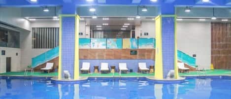 Una piscina interior con múltiples carriles, sillones de descanso y una pared de azulejos con un mural.
