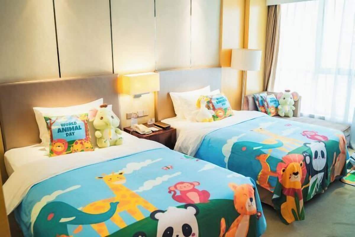 Habitación de hotel con dos camas, cada una con una colcha de colores con motivos de animales. Hay peluches en las camas, una mesita de noche con una lámpara y una ventana con cortinas.