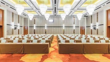 A conference room with rows of tables and chairs, large chandeliers, and a patterned carpet.