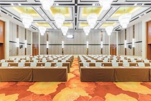 Un salón de conferencias con filas de mesas y sillas, grandes candelabros y una alfombra estampada.