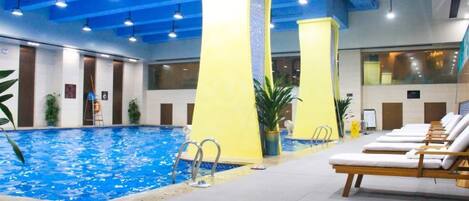 An indoor swimming pool with a blue water feature, surrounded by lounge chairs and potted plants.