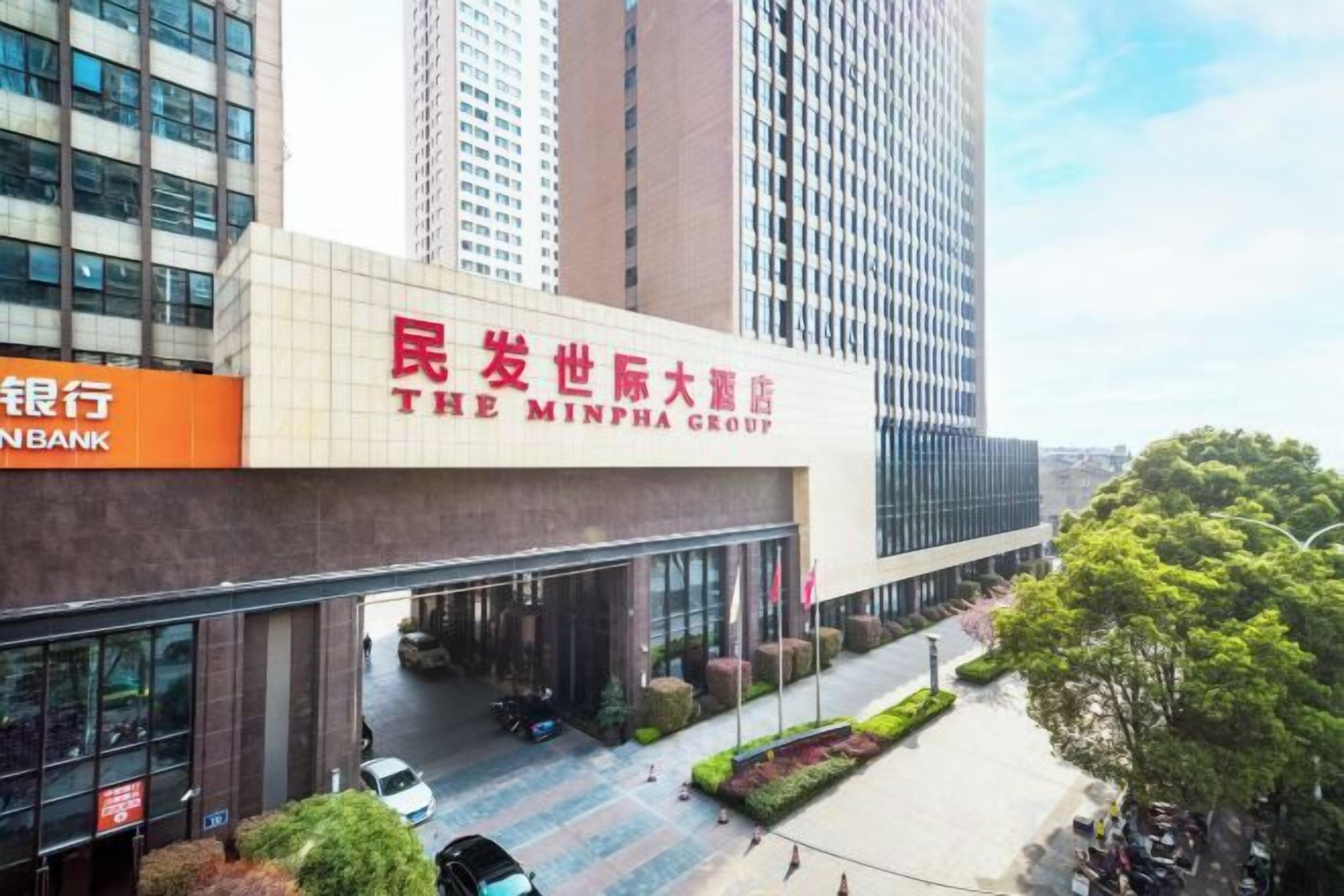 Un hotel con fachada moderna, un llamativo letrero de 'The Minpha Group' y una entrada visible con zona de estacionamiento.