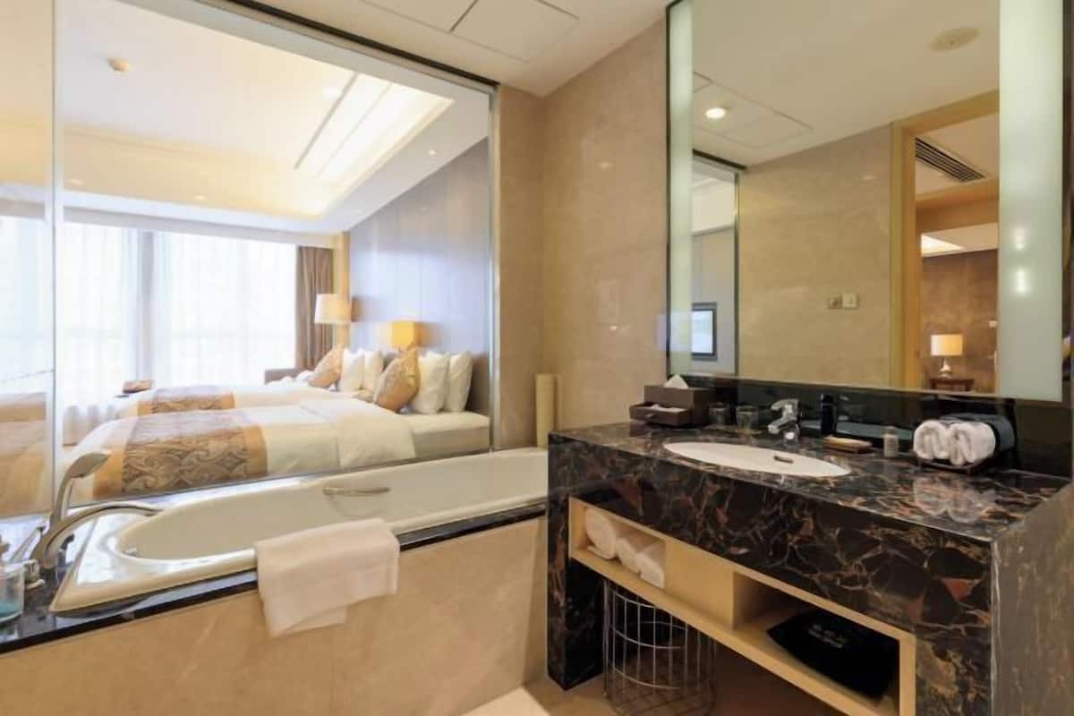Un baño de hotel con bañera, lavamanos, espejo y una cama visible a través de un panel de vidrio.