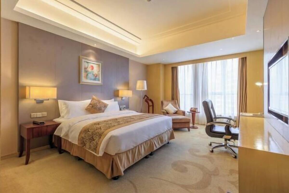 Habitación de hotel con una cama grande, dos mesitas de noche con lámparas, un escritorio con silla y un ventanal con cortinas.