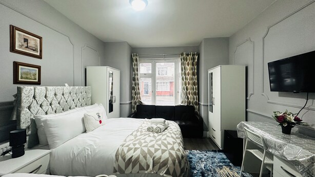 A hotel room with a bed, a desk, a chair, a wardrobe, a television, and a window with curtains. - Heathrow roomzzzz B&B (Hayes)
