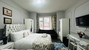 A hotel room with a bed, a desk, a chair, a wardrobe, a television, and a window with curtains. - Heathrow roomzzzz B&B (Hayes)