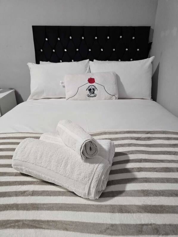 Una cama bien tendida con ropa blanca y una colcha a rayas, un almohadón con diseño de rosa y una toalla enrollada sobre la cama.