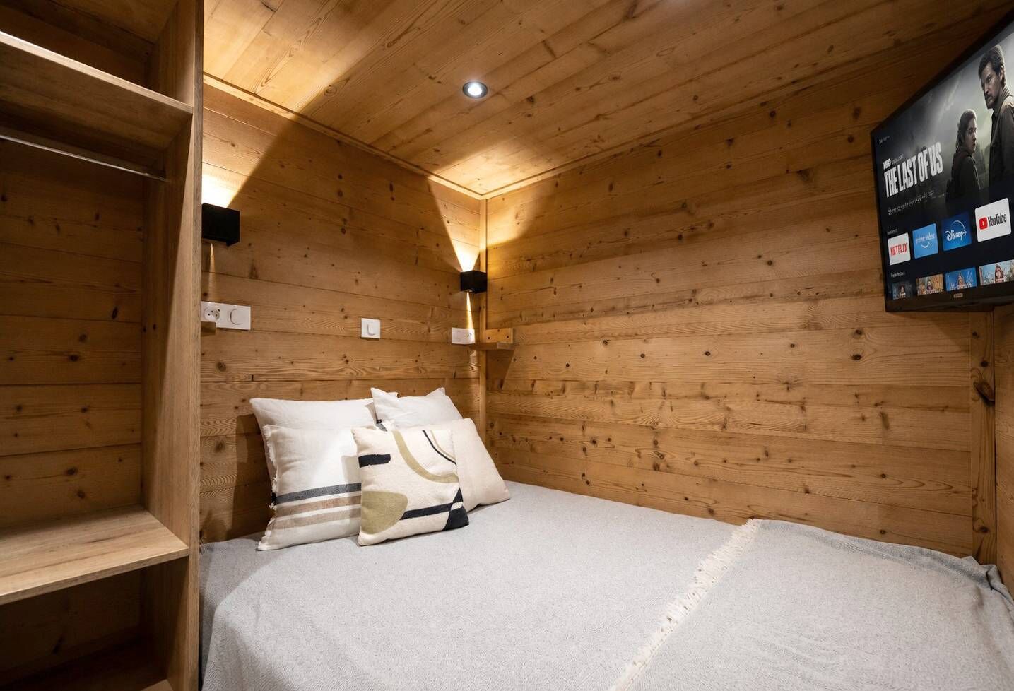 A wooden cabin room with a bed, pillows, a wall-mounted TV, and a shelf.