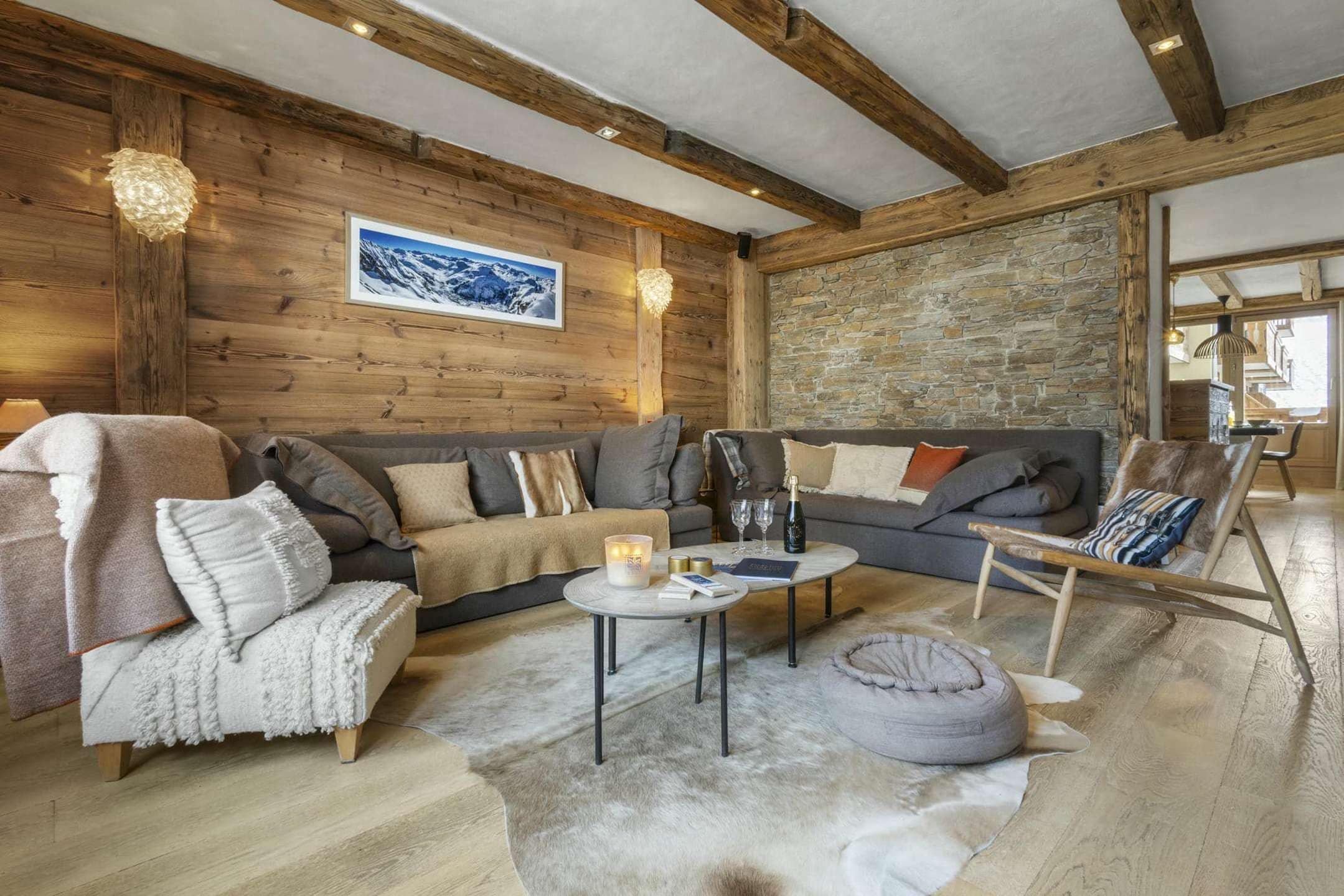 A cozy living room with a wooden ceiling, stone wall, and a large window. There is a sofa, a chair, and a coffee table with candles and books.