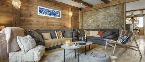 A cozy living room with a wooden ceiling, stone wall, and a large window. There is a sofa, a chair, and a coffee table with candles and books.