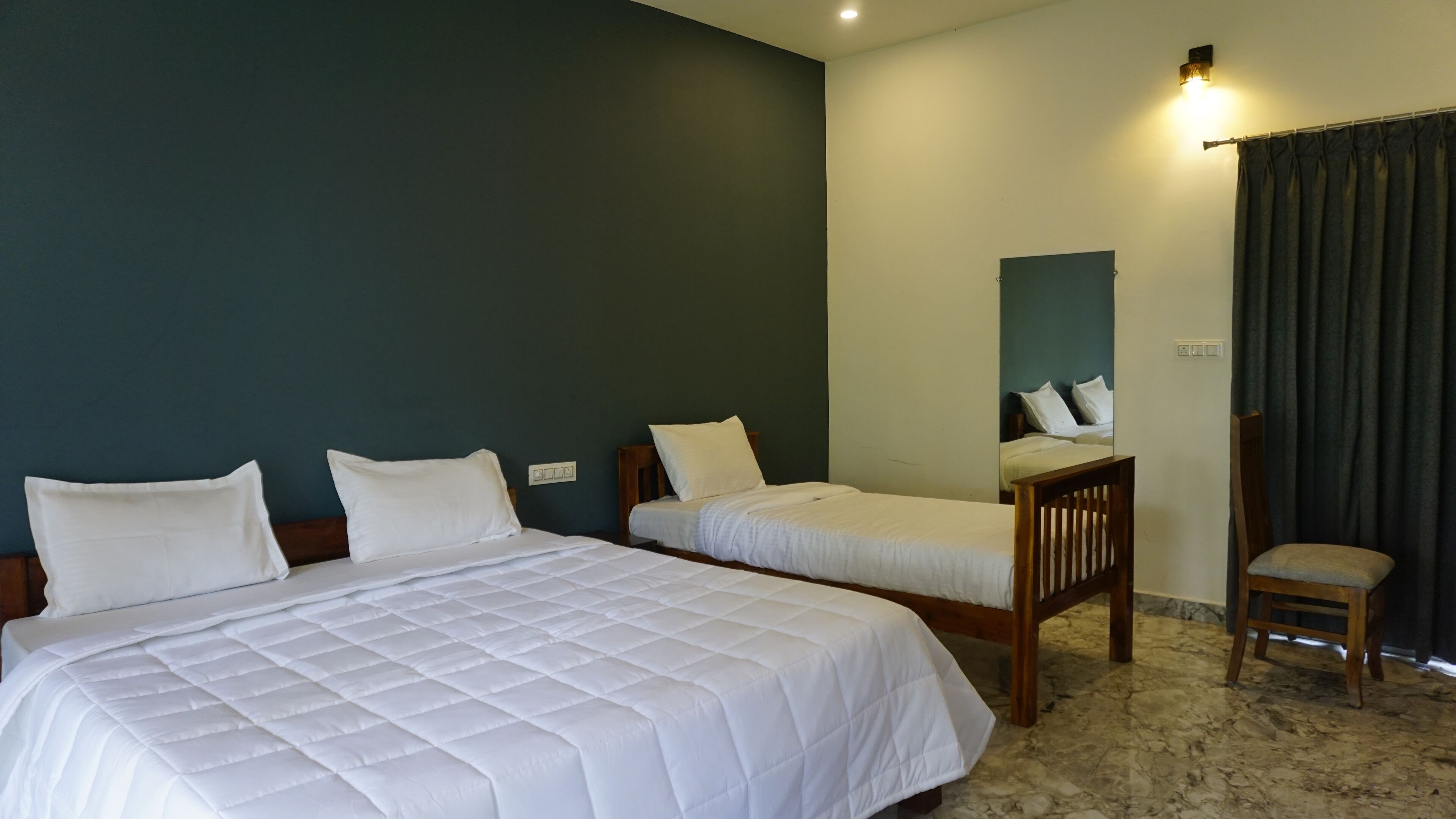 A hotel room with two single beds, a wooden chair, a dark curtain, and a wall-mounted light fixture.