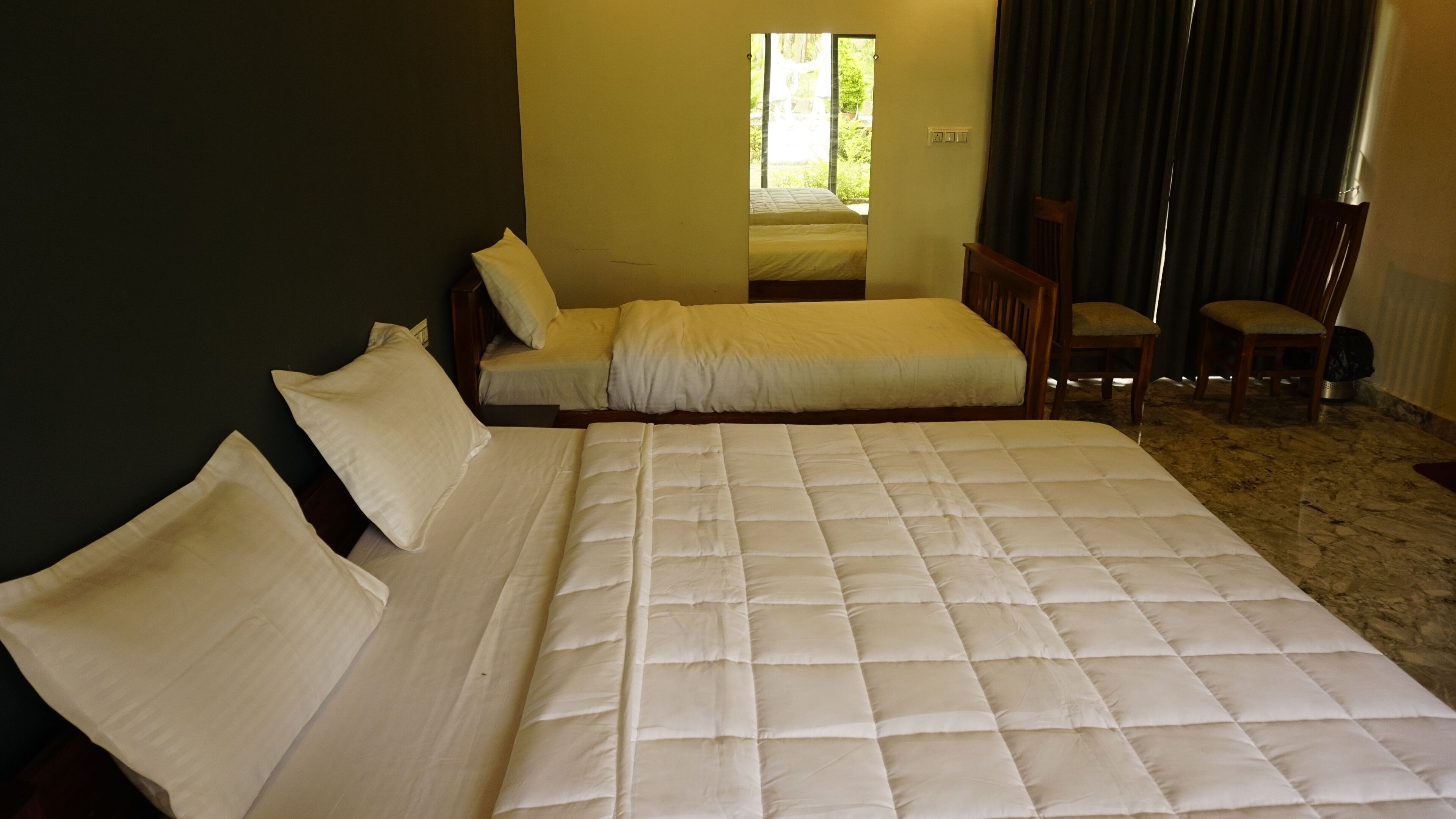 A room with a large bed, a smaller bed, two chairs, and a window with a view of greenery.