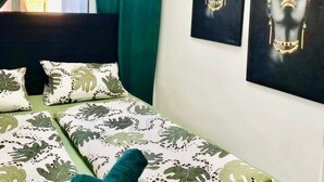 Un lit bien fait, recouvert d’une couvre-lit à motifs de feuilles, avec des rideaux verts et deux œuvres d’art accrochées au mur.