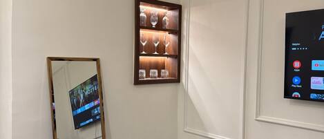 A room with a TV mounted on the wall, a wooden shelf with glassware, a mirror, and a black trash bin.