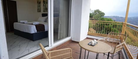 Un balcón con sillas de mimbre y una mesa, un dormitorio con cama y puerta corrediza de vidrio, y vistas a zonas verdes y colinas.