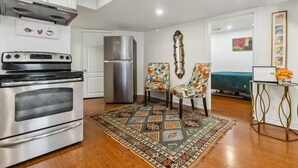 Cocina compacta con horno de acero inoxidable, refrigerador y armarios blancos. Hay una alfombra estampada sobre el suelo de madera, y dos sillas con motivos florales.
