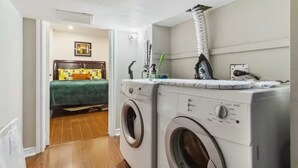 Un lavadero pequeño con dos lavadoras de carga frontal, una cama con colcha verde y una mesita de noche con lámpara.