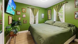 A bedroom with a green wall, a bed with a green bedspread, a television on the wall, and several framed pictures. - Coastal Comfort 2BR Getaway St Pete (St. Petersburg)