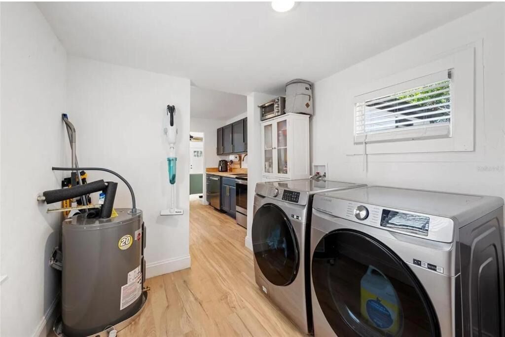 A laundry area with two washing machines, a water heater, and a window with blinds.