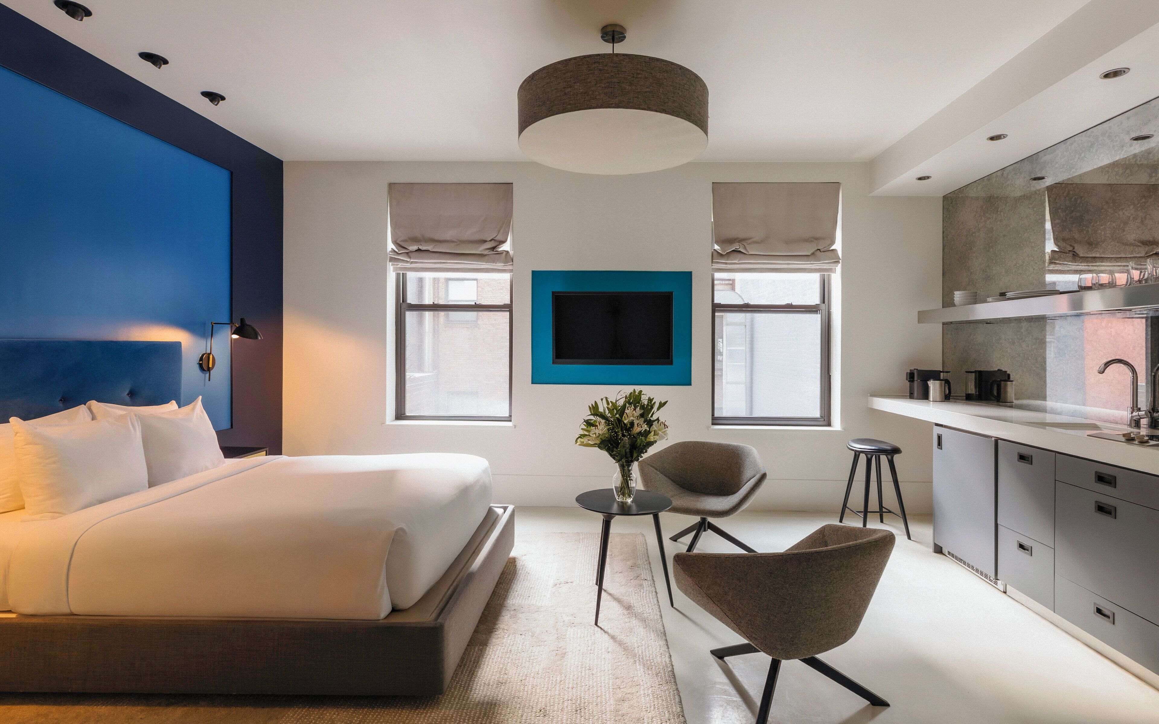 A modern bedroom with a large bed, a sitting area with a chair, a small table, and a kitchen area with a sink and cabinets.