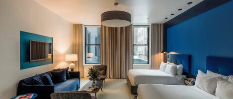Ein Hotelzimmer mit zwei Betten, einem Schlafsofa, einem kleinen Tisch, einem Fernseher und einem Fenster mit Vorhängen.