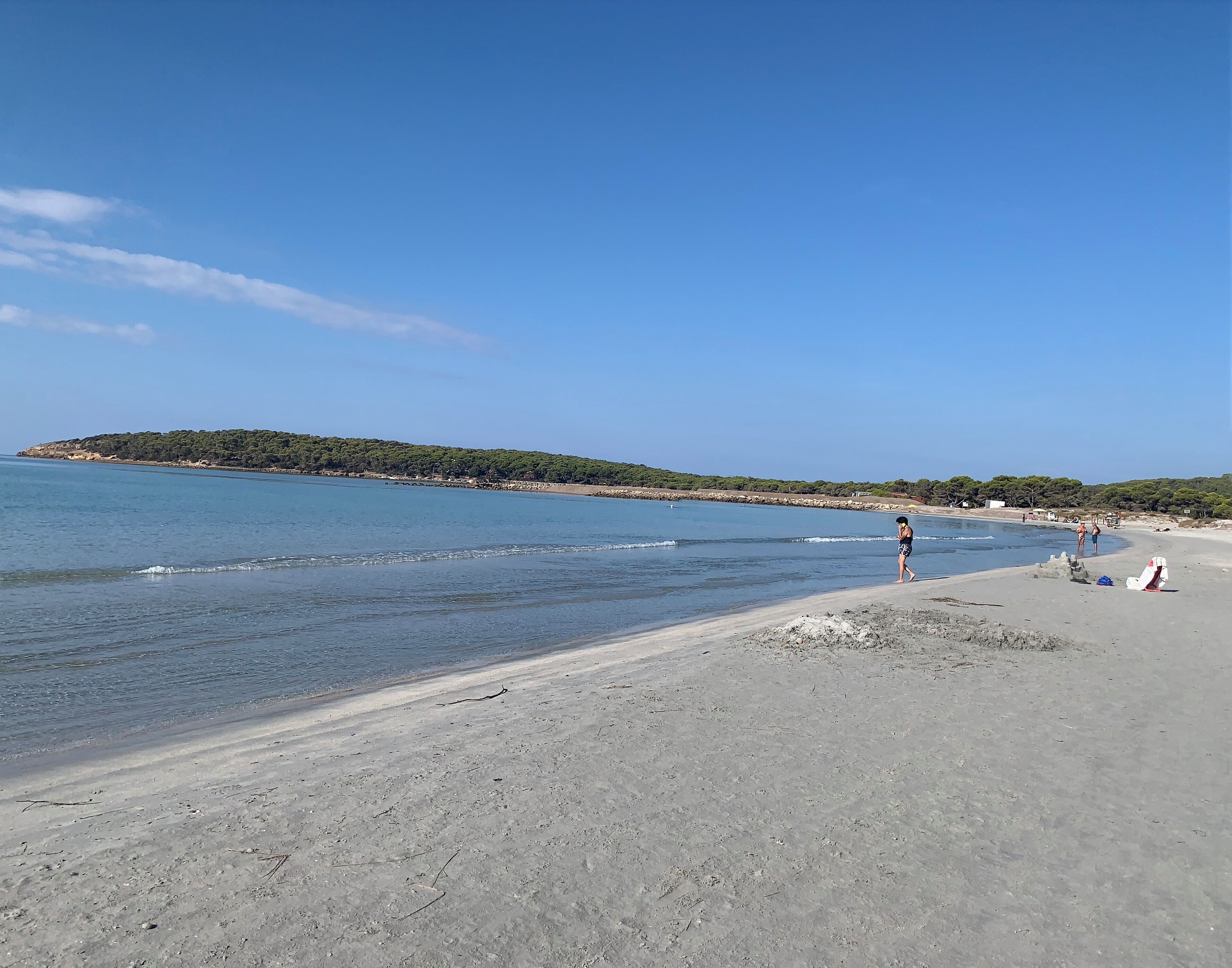 Une plage à l’eau d’un bleu limpide, bordée d’un rivage sablonneux, avec une colline verdoyante qui se dessine à l’horizon.