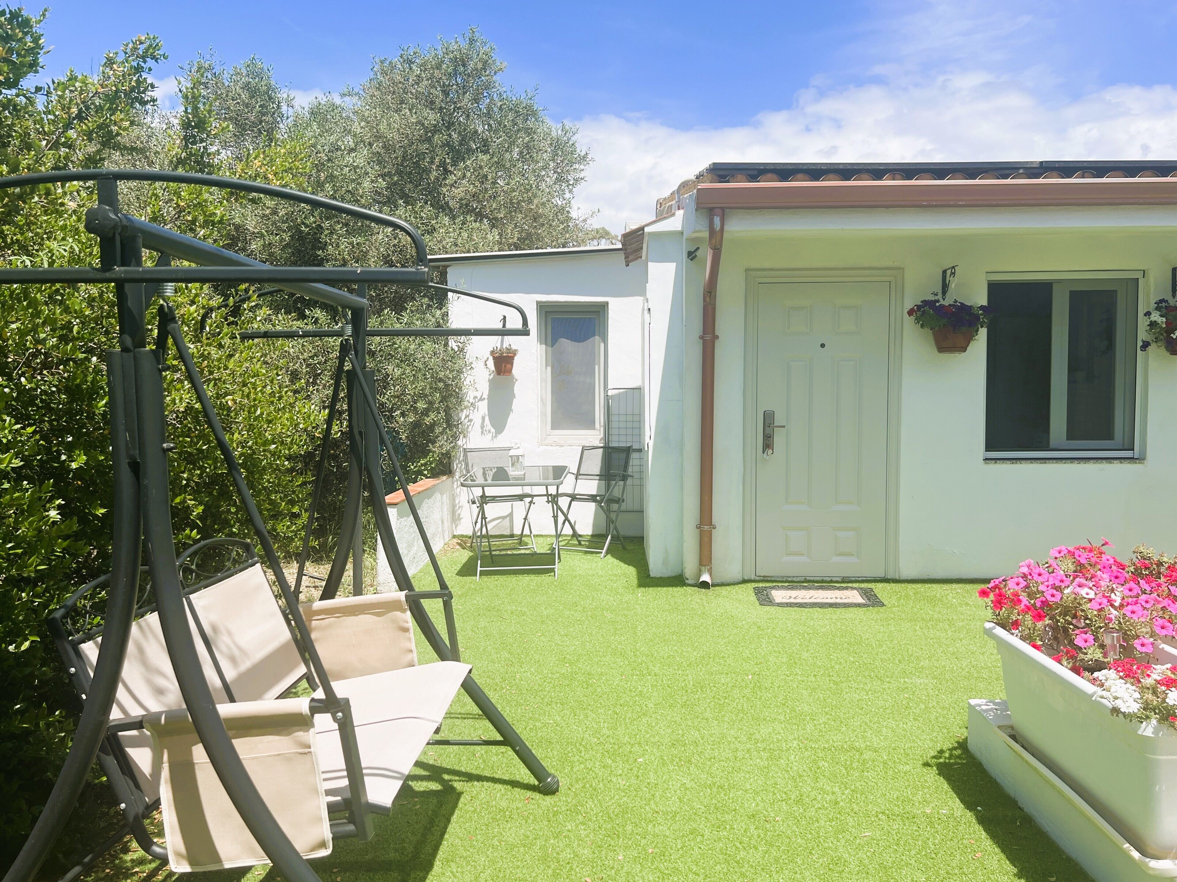 Eine Terrasse mit Schaukel und einer weißen Tür, sowie ein kleiner Garten mit Blumen.