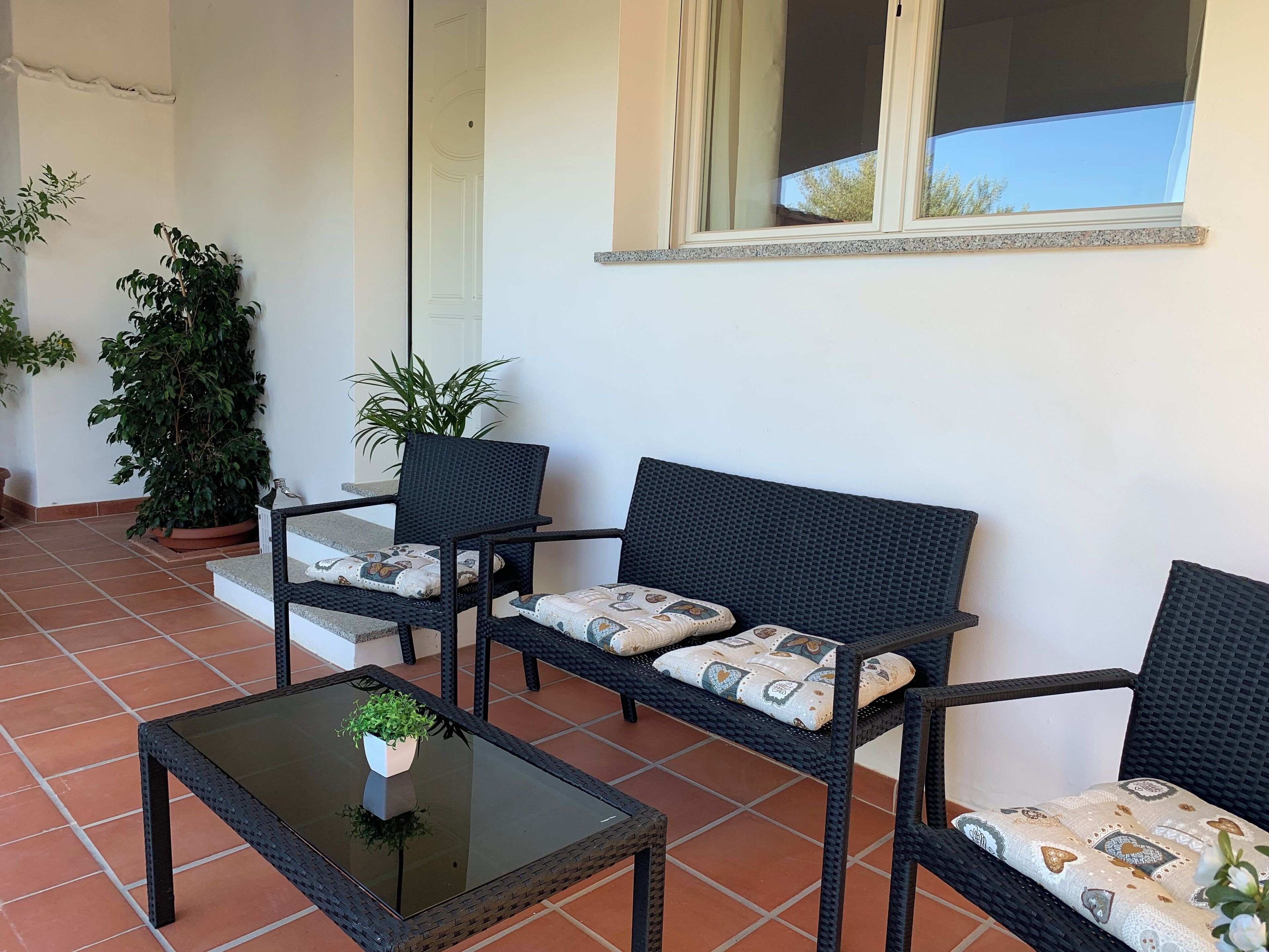 A patio with a glass-top table, two black wicker chairs with patterned cushions, and a potted plant.
