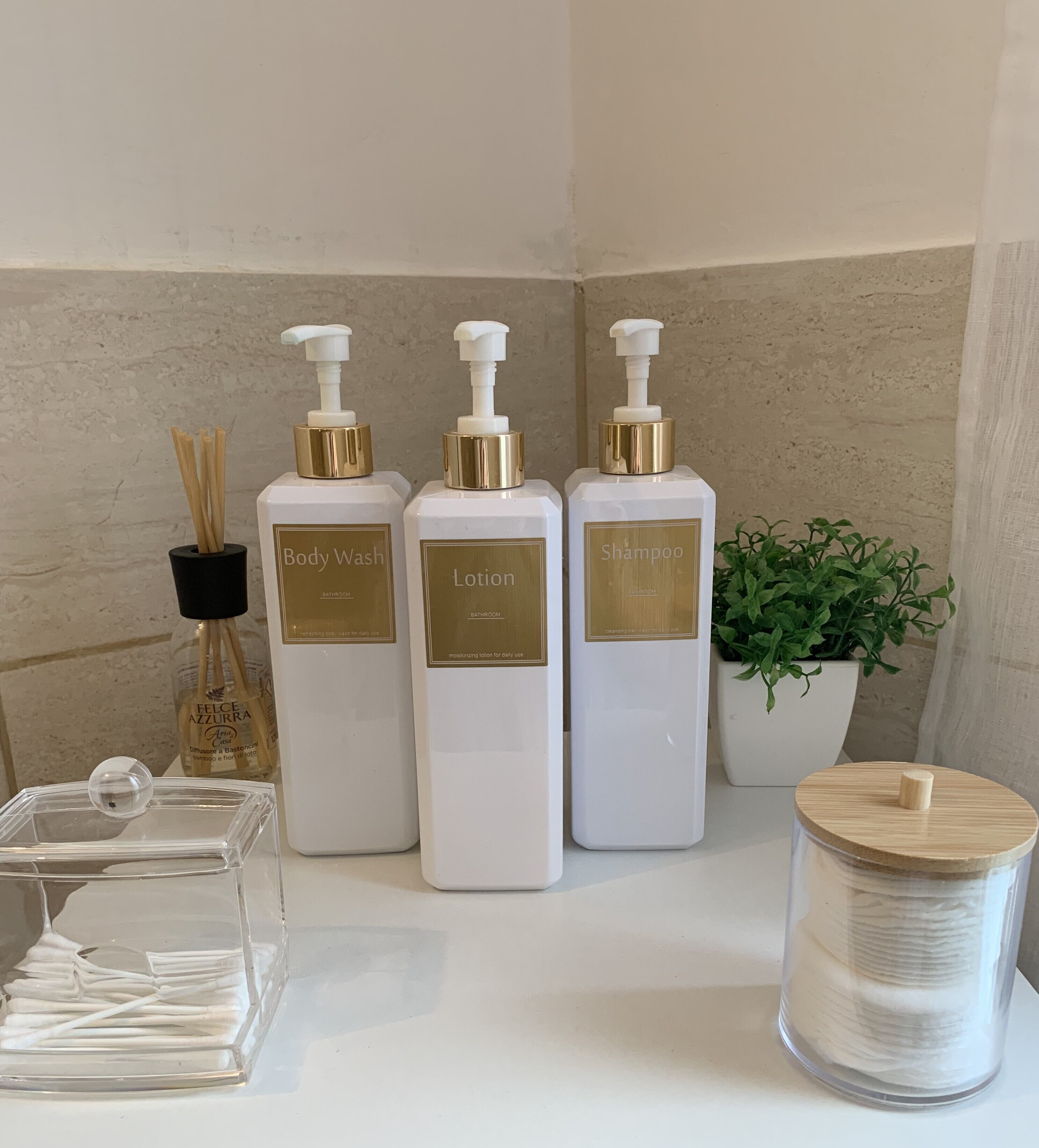 Tres botellas blancas con etiquetas doradas que dicen "Body Wash", "Lotion" y "Shampoo" sobre una encimera de baño.